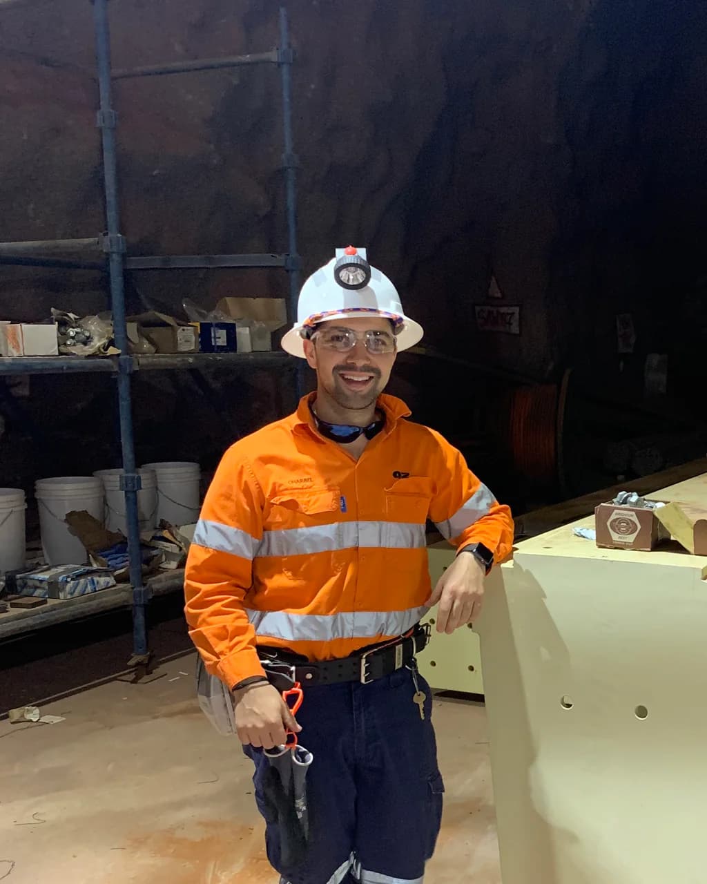 Charbel Fata, Founder & CEO of Tracta, underground in a hard hat with a cap lamp and hi-vis workwear, leaning on a large steel beam.
